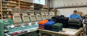 Folded stacks of blank polo shirts and jackets next to a Tajima embroidery machine at Northwest Custom Apparel Milton facility