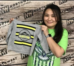 A smiling person holding up a gray sweatshirt with a screen-printed design. The design includes a graphic element with three horizontal stripes and the word "FLAGSTONE" prominently displayed in the center. The person is standing in front of a backdrop with text and logo graphics, which appears to be the branding wall of a custom apparel company. The overall image conveys a cheerful and professional atmosphere, often associated with small businesses showcasing their custom printing services.