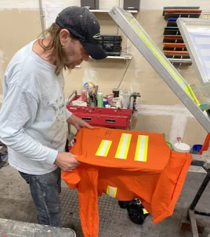 A person in a workshop setting, focused on a screen printing task. They are wearing a cap and a long-sleeved shirt, both splattered with paint, suggesting that they are engaged in a creative or manual job. In front of the individual is a bright orange safety vest with reflective stripes being held out for inspection or preparation, likely to be screen printed or customized. The workshop has various tools and equipment, hinting at a space where hands-on work is performed