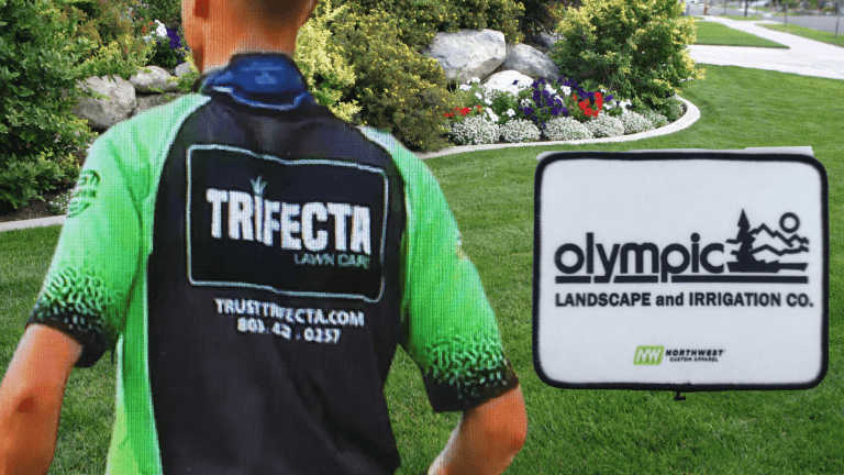 A landscaper in a vibrant green and black uniform branded with 'TRIFECTA LAWN CARE' and the website 'trusttrifecta.com' stands with their back to the camera, overlooking a well-manicured lawn with a sign reading 'Olympic Landscape and Irrigation Co.', indicating a professional landscaping service