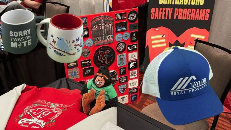 A variety of budget-friendly promotional items displayed on a table, including a red WSU Cougar Jacket, a blue and white trucker cap with a logo, a custom mug, assorted pins on a blackboard, and a small plush Sasquatch toy, next to a poster about safety apparel programs.