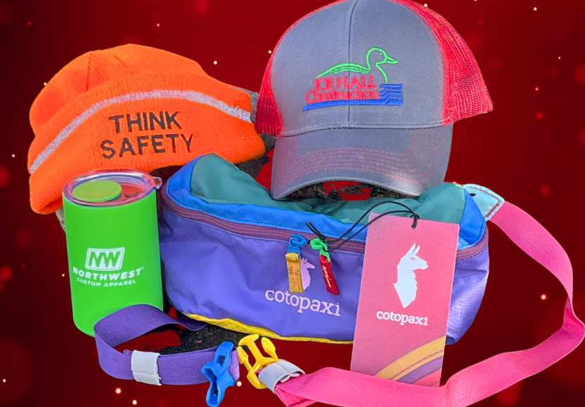 Assortment of branded items including a bright orange "THINK SAFETY" beanie, a "Joe Hail Construction" cap, a green "Northwest Custom Apparel" tumbler, and a colorful "Cotopaxi" sling bag against a sparkly red backdrop.